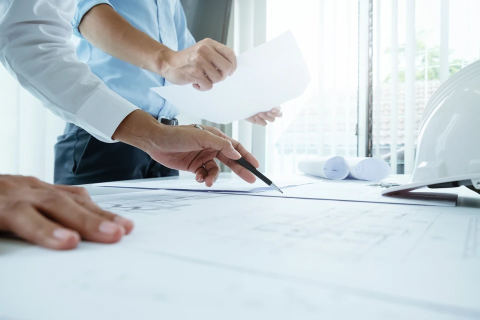 Personne en chemise à carreaux travaillant sur des plans architecturaux posés sur une table en bois, tenant un stylo et analysant des documents techniques